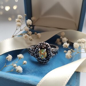 May include: A silver ring with a floral design, featuring a central, iridescent gemstone. The ring is displayed in a blue velvet ring box, accented with white flowers and a cream ribbon.