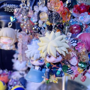 以下が含まれることがあります： アニメスタイルのキャラクターキーホルダーのコレクション。ツンツンしたブロンドの髪や黒髪のフィギュア、カラフルなビーズが特徴です。左上隅にAris Studioのロゴが見えます。チャームには小さなフィギュアや円形のレジンパーツが含まれています。