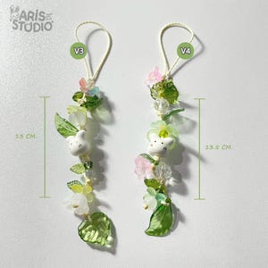 Puede incluir: Dos colgantes de cuentas, uno de 13 cm y otro de 13,5 cm de largo, con figuras de conejos blancos, hojas verdes y flores rosas y blancas. Los colgantes están ensartados en un cordón color crema.