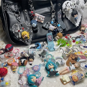 Può includere: Una borsa in similpelle nera decorata con numerosi portachiavi a tema anime, tra cui figurine in miniatura, ali d'angelo, telefoni a conchiglia e palloni sportivi. La borsa presenta un'etichetta Aris Studio e vari ciondoli colorati sparsi.