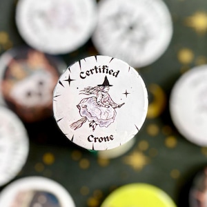 Op de afbeelding: Een verzameling ronde buttons met verschillende ontwerpen, waaronder een heks op een bezem met de tekst "Certified Crone". Andere buttons tonen hemelse en abstracte illustraties. De buttons zijn gerangschikt op een donkergroen oppervlak met gouden ster- en maanaccenten.