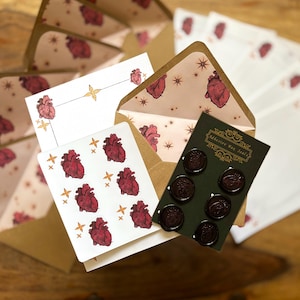 May include: A stationery set featuring white envelopes and paper with a red anatomical heart and gold star pattern. A dark green card displays six circular dark red adhesive wax seals with heart imprints and the text Adhesive Wax Seals.