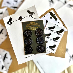 May include: A selection of raven-themed stationery. The image shows black wax seals, stickers, and cards adorned with raven and branch illustrations. The packaging displays the text "Adhesive Wax Seals".