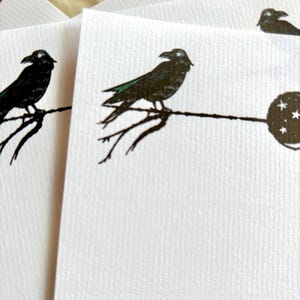 May include: White stationery cards featuring a black crow perched on a branch design. The crow has green accents on its wings. A full moon with stars is also visible. The cards have a textured surface.