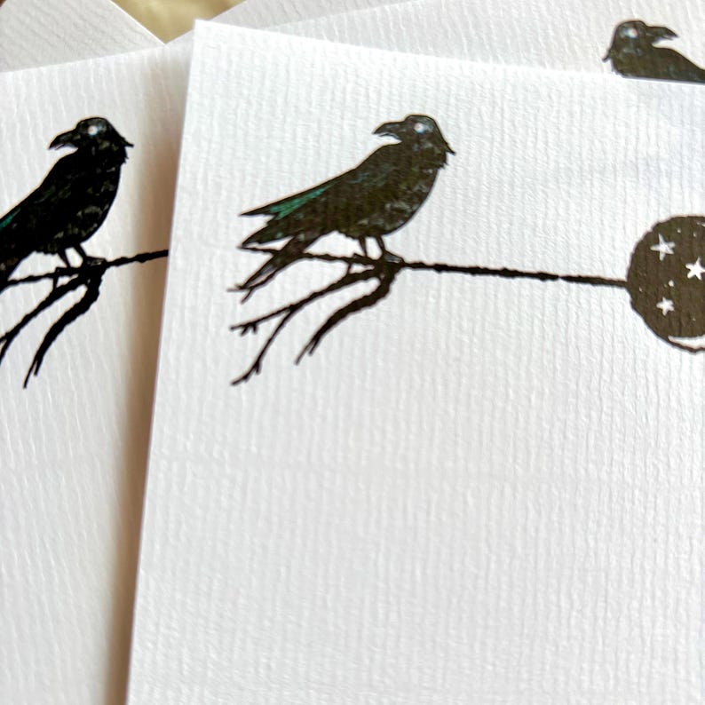 May include: White stationery cards featuring a black crow perched on a branch design. The crow has green accents on its wings. A full moon with stars is also visible. The cards have a textured surface.