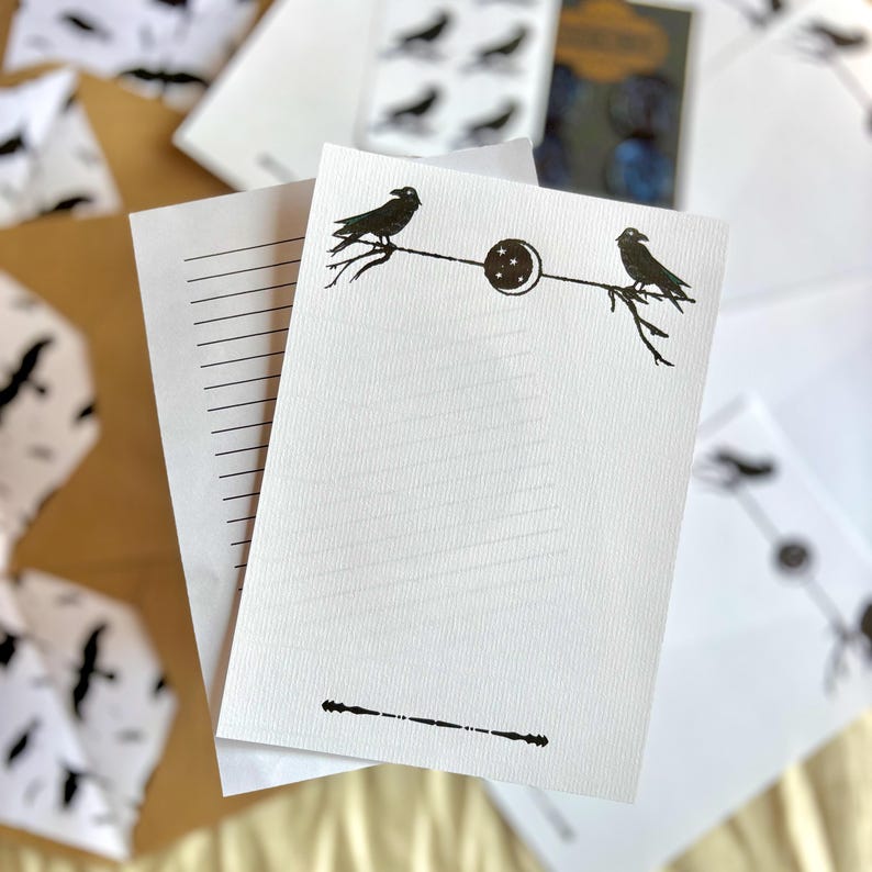 May include: White stationery set with a raven and moon design. The set includes lined paper and envelopes with a black raven illustration perched on a branch. The stationery is suitable for writing letters or notes.