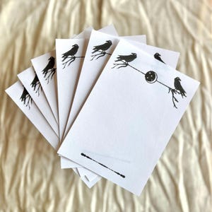 May include: A collection of white writing paper featuring black raven designs. Each sheet has a raven perched on a branch, with a crescent moon and star motif on some. The paper is fanned out, displaying the illustrations.