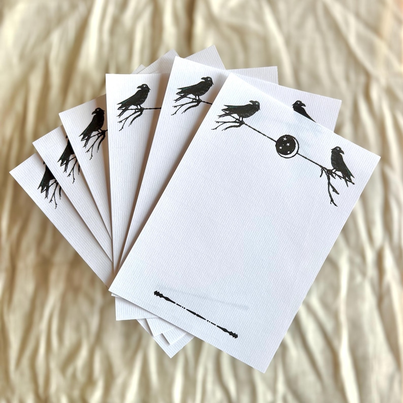 May include: A collection of white writing paper featuring black raven designs. Each sheet has a raven perched on a branch, with a crescent moon and star motif on some. The paper is fanned out, displaying the illustrations.