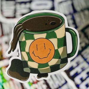 May include: A sticker of a coffee mug with a green and white chequered pattern. The mug is overflowing with dark brown coffee, and has an orange smiley face on the front. The mug has a green handle and rim.