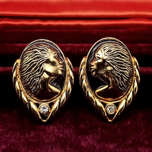 May include: A pair of gold-toned earrings. Each earring features an oval cameo with a profile of a woman's face, set against a black background. The earrings are framed by a gold leaf design and a small, clear crystal at the bottom.