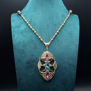 May include: Gold-toned necklace with a pendant featuring an oval filigree design. The pendant is adorned with pink, blue, and purple gemstones. The necklace is displayed on a teal velvet bust, against a dark background.