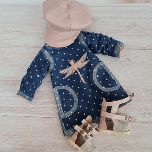 May include: A doll's denim dress with white polka dots, a pink dragonfly design, and matching pink sandals. A pink cap sits above the dress. The dress has two front pockets. The sandals have a textured fabric.