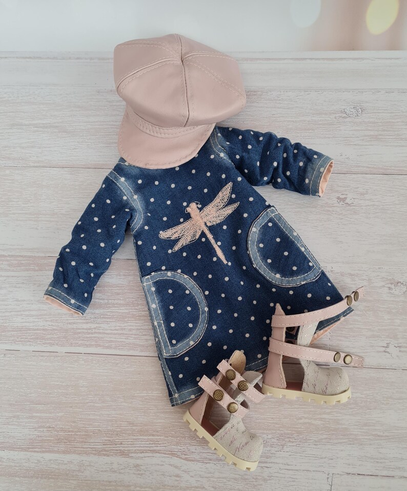 May include: A doll's denim dress with white polka dots, a pink dragonfly design, and matching pink sandals. A pink cap sits above the dress. The dress has two front pockets. The sandals have a textured fabric.