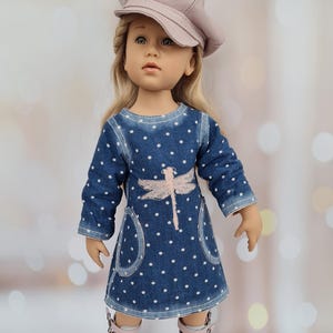 May include: A doll wearing a denim dress with white polka dots and a pink dragonfly design. The doll has blonde hair and is wearing a pink cap and white and pink shoes. The background is blurred with bokeh lights.