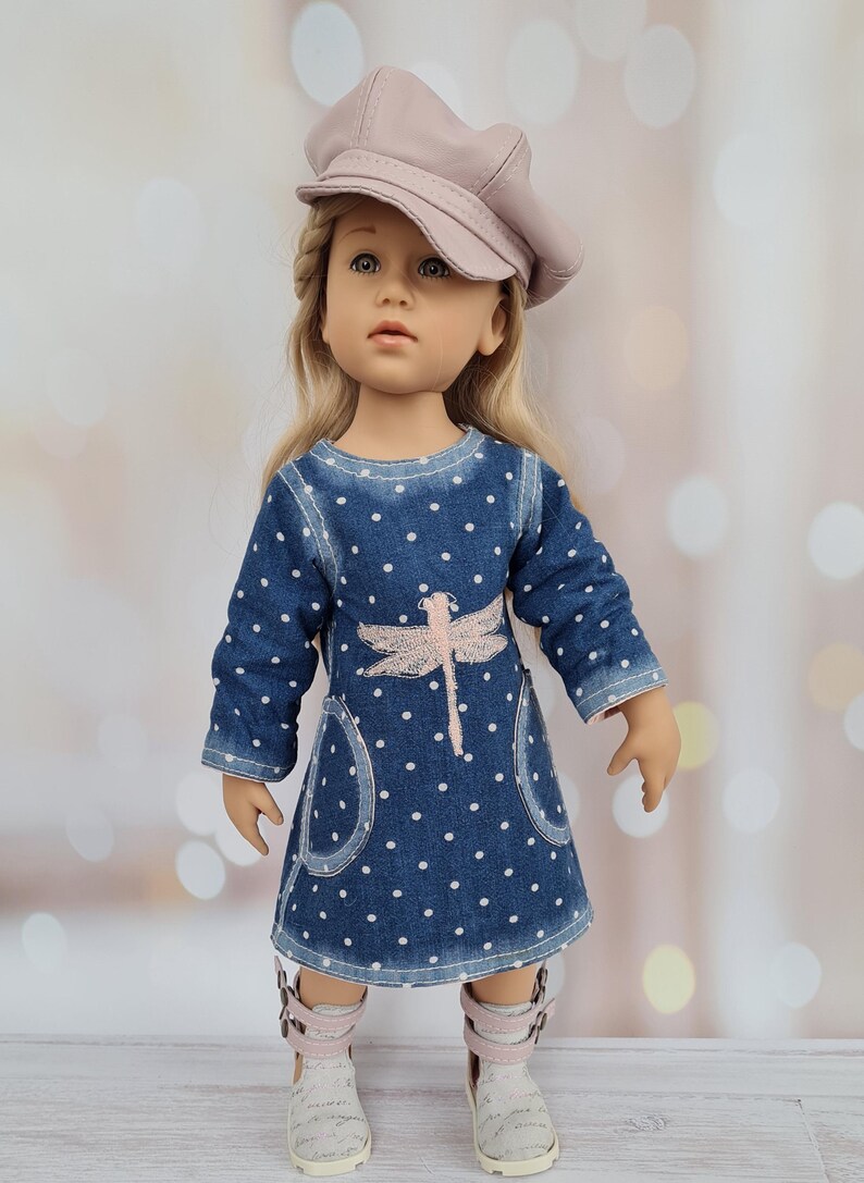 May include: A doll wearing a denim dress with white polka dots and a pink dragonfly design. The doll has blonde hair and is wearing a pink cap and white and pink shoes. The background is blurred with bokeh lights.
