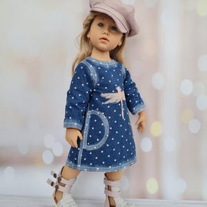 May include: A doll wearing a blue denim dress with white polka dots and a pink dragonfly design. The doll is also wearing a pink cap and white and pink sandals. The doll has blonde hair and is standing in front of a blurred background.