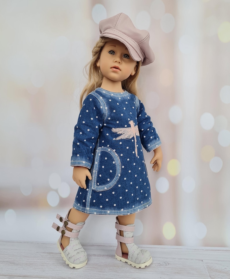 May include: A doll wearing a blue denim dress with white polka dots and a pink dragonfly design. The doll is also wearing a pink cap and white and pink sandals. The doll has blonde hair and is standing in front of a blurred background.