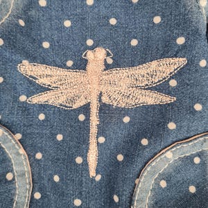 May include: Close-up of a denim fabric with white polka dots and a light pink embroidered dragonfly. The denim has a pocket detail with a light pink trim. The dragonfly is centered on the fabric.