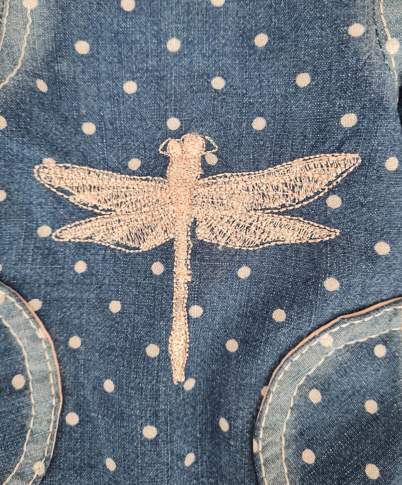 May include: Close-up of a denim fabric with white polka dots and a light pink embroidered dragonfly. The denim has a pocket detail with a light pink trim. The dragonfly is centered on the fabric.