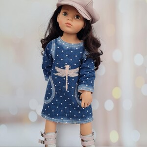 May include: A doll wearing a blue denim dress with white polka dots and a pink dragonfly design. The doll has long dark hair and a light pink cap. She is wearing gray and pink boots with buckles.