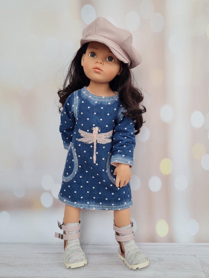 May include: A doll wearing a blue denim dress with white polka dots and a pink dragonfly design. The doll has long dark hair and a light pink cap. She is wearing gray and pink boots with buckles.