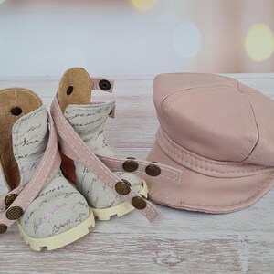 May include: A pair of doll-sized ankle boots and a matching hat. The boots are off-white with script writing, pink straps, and bronze-colored studs. The hat is a solid, light pink, and the items are displayed on a light wood surface.