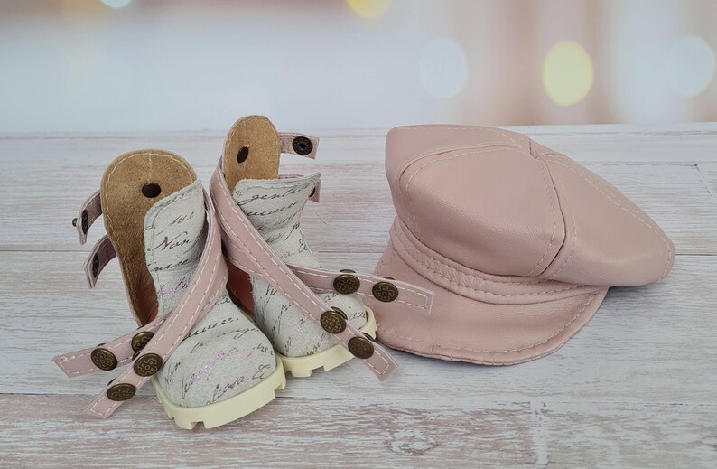 May include: A pair of doll-sized ankle boots and a matching hat. The boots are off-white with script writing, pink straps, and bronze-colored studs. The hat is a solid, light pink, and the items are displayed on a light wood surface.
