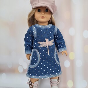 May include: A doll wearing a denim dress with white polka dots and a pink dragonfly design. The doll also wears a matching pink cap and white and pink sandals. The doll has blonde hair and blue eyes.