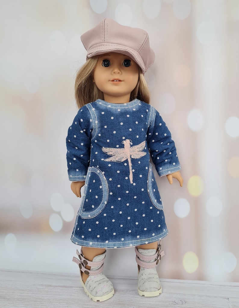 May include: A doll wearing a denim dress with white polka dots and a pink dragonfly design. The doll also wears a matching pink cap and white and pink sandals. The doll has blonde hair and blue eyes.