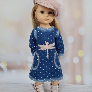 May include: A doll wearing a blue denim dress with white polka dots and a pink dragonfly design. The dress has pockets and is paired with white sandals with pink straps and a pink cap. The doll has blonde hair and blue eyes.