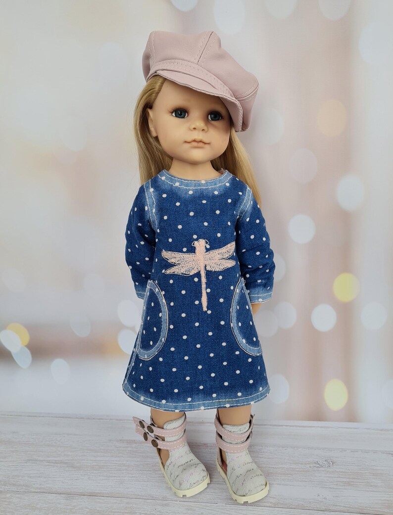 May include: A doll wearing a blue denim dress with white polka dots and a pink dragonfly design. The dress has pockets and is paired with white sandals with pink straps and a pink cap. The doll has blonde hair and blue eyes.