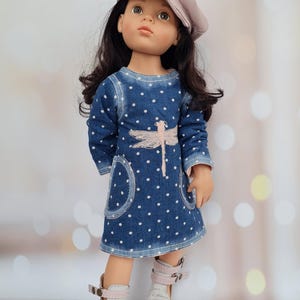 May include: A doll wearing a denim dress with white polka dots and a pink dragonfly design. The doll has dark hair and is wearing a light pink cap and white and pink sandals.