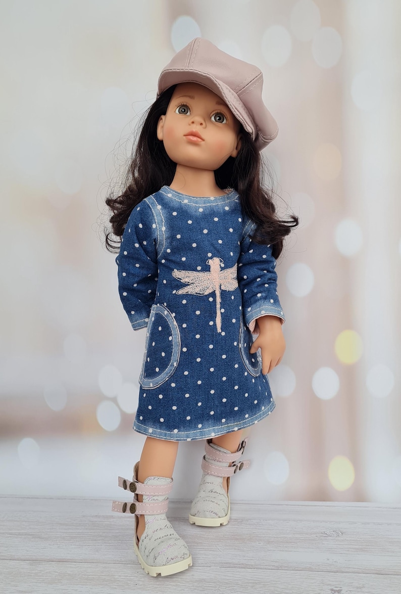 May include: A doll wearing a denim dress with white polka dots and a pink dragonfly design. The doll has dark hair and is wearing a light pink cap and white and pink sandals.