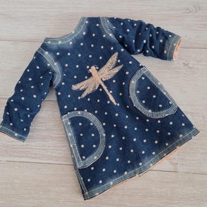 May include: A blue denim dress with white polka dots and a light pink dragonfly applique. The dress has long sleeves, two front pockets, and light pink trim. The dress is designed for a child.