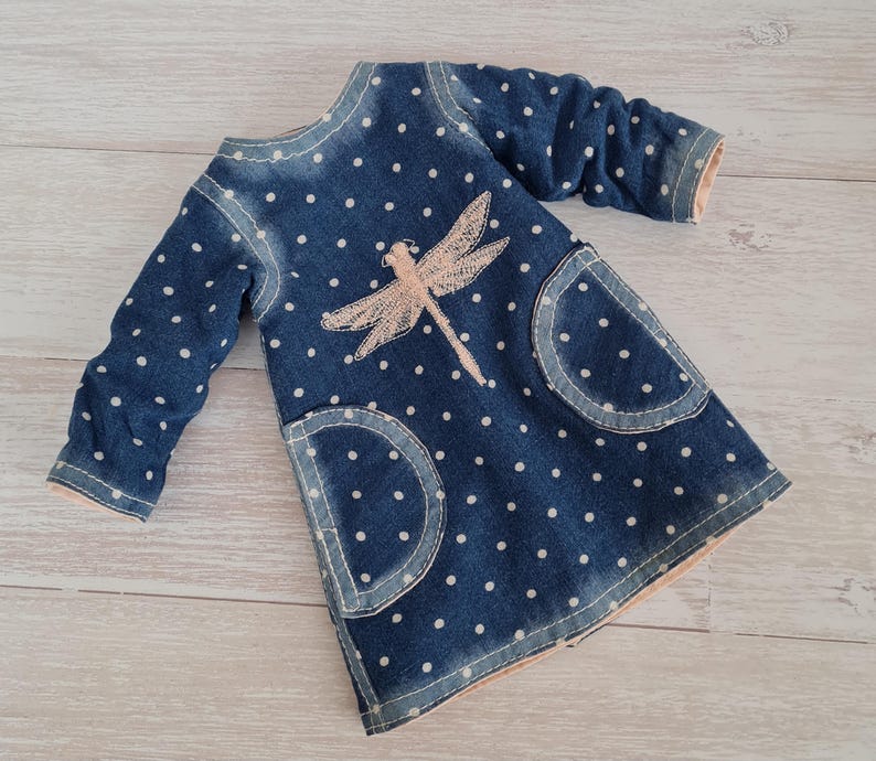 May include: A blue denim dress with white polka dots and a light pink dragonfly applique. The dress has long sleeves, two front pockets, and light pink trim. The dress is designed for a child.