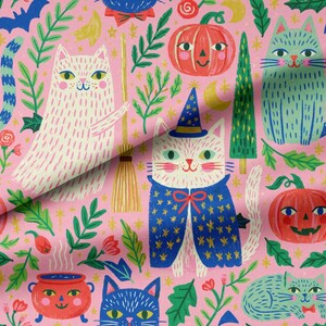 Puede incluir: Una tela rosa con un patrón repetitivo de gatos, calabazas y escobas. Los gatos aparecen en blanco, azul y verde, algunos con sombreros de mago y capas azules. Calabazas naranjas con caras, árboles verdes y pequeñas flores están dispersas por todo el diseño.