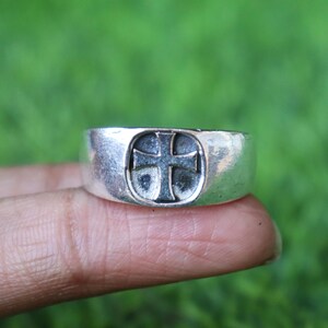 May include: A silver ring featuring a raised cross design. The ring is wide and has a slightly textured surface. The cross is centrally placed and has a dark, recessed background.
