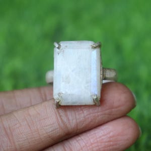 May include: A silver ring featuring a large, rectangular, iridescent, off-white gemstone. The gemstone is set with four prongs. The ring is held against a blurred green background.
