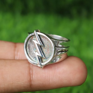 Könnte beinhalten: Ein silberner Ring mit einem Blitzsymbol. Der Ring wird an einem Finger getragen und hat eine polierte Oberfläche. Der Hintergrund ist ein verschwommenes Grün. Das Design des Rings ist kreisförmig.