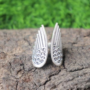 Puede incluir: Un anillo plateado con un diseño abierto que presenta dos formas de alas estilizadas. Las alas tienen patrones de plumas detallados y un acabado texturizado. El anillo está sobre un fondo de madera natural.