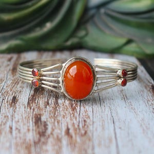 Peut inclure: Bracelet manchette argenté avec une pierre précieuse ovale orange au centre. Le bracelet comporte plusieurs bandes parallèles et est orné de petites pierres précieuses rouges rondes de chaque côté de la pierre centrale. Le bracelet est présenté sur une surface en bois.