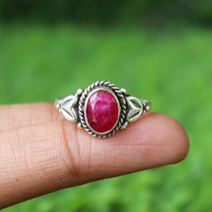 May include: A silver ring featuring an oval, deep red gemstone. The gemstone is set in a decorative, rope-like border. The ring has stylized triangular accents on either side of the gemstone, creating a unique and elegant design.
