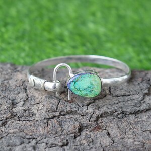 May include: A silver-toned bracelet with a hook closure and a large, oval-shaped, green gemstone. The gemstone has blue and purple accents. The bracelet is displayed on a textured, brown surface with a green background.