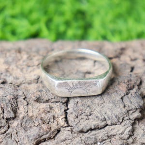 Può includere: Anello d'argento con una faccia rettangolare con un disegno a raggiera. L'anello è posto su una superficie strutturata marrone scuro, con uno sfondo verde sfocato.