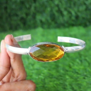 Op de afbeelding: Een zilveren manchetarmband met een grote, facetgeslepen, ovaalvormige gele edelsteen. De armband heeft een eenvoudig, open ontwerp met de edelsteen gezet in een zilveren bezel. De achtergrond is een wazig groen.