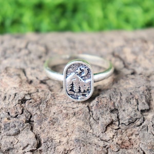 Può includere: Anello d'argento con un ciondolo rettangolare raffigurante un paesaggio montano e boschivo, con luna e stella. L'anello è posto su una superficie marrone strutturata.