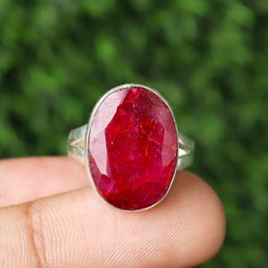 Op de afbeelding: Een zilveren ring met een grote, ovaal geslepen robijn. De robijn heeft een diepe, levendige rode kleur en is gezet in een eenvoudige, elegante zilveren band. De ring wordt getoond tegen een vage groene achtergrond.