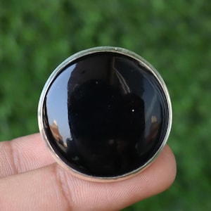 May include: A round, black gemstone ring with a silver-colored bezel. The polished black stone reflects light, creating a glossy appearance. The ring is held by a person's fingers against a blurred green background.