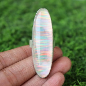 May include: A long, oval-shaped ring with a large, iridescent opal stone set in a silver band. The opal displays a play of color with hues of blue, green, and orange. The ring is held against a green background.