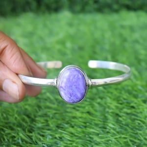 Pode incluir: Uma pulseira de punho prateada com uma grande pedra ovalada de cor lavanda. A pedra tem uma aparência marmorizada com veios brancos. A pulseira é apresentada sobre um fundo de relva verde.